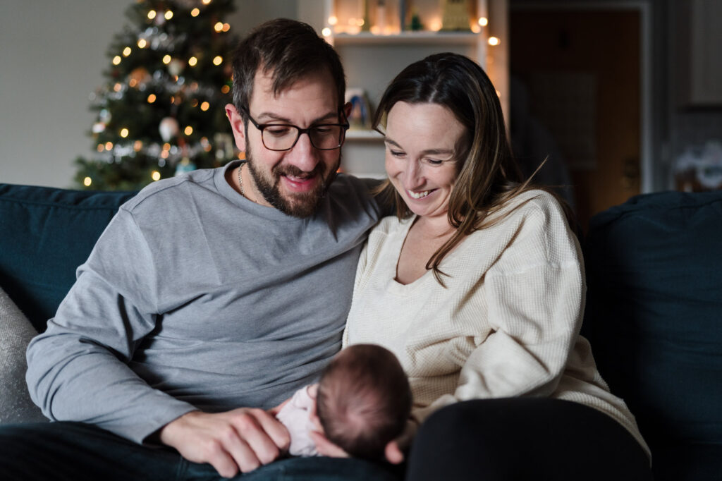 Parents are holding a newborn with happy faces on before the Christmas tree at home during a newborn photography session by Ivy Lulu Photography